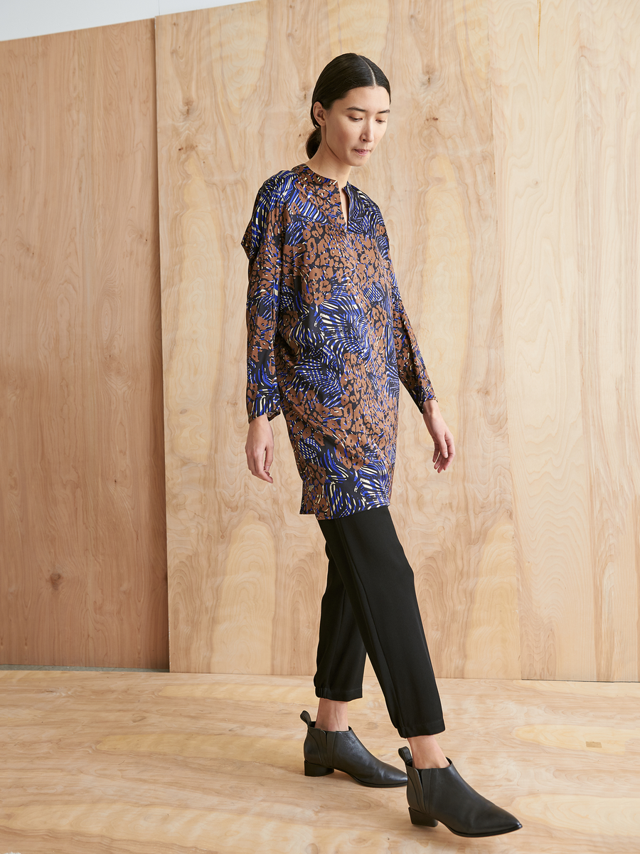 A woman with dark hair wears the Zero + Maria Cornejo Long-Sleeved Hex Tunic Dress in blue and brown cheetah print, styled with black pants and ankle boots, standing indoors before a light wood panel wall on a wooden floor.