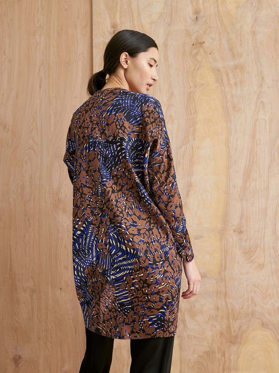 A woman with dark hair tied back stands sideways, facing away from the camera, wearing the Zero + Maria Cornejo Long-Sleeved Hex Tunic Dress with brown, blue, and black abstract designs, set against a light wooden background.