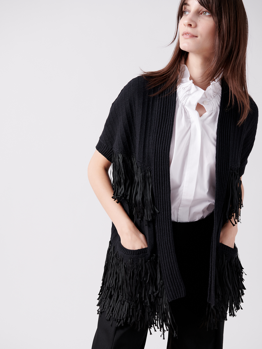 A woman with long brown hair stands against a light gray background, looking slightly right. She wears the Fringe Hex Shrug by Zero + Maria Cornejo over a white blouse and black pants, with hands tucked in the shrug's pockets.