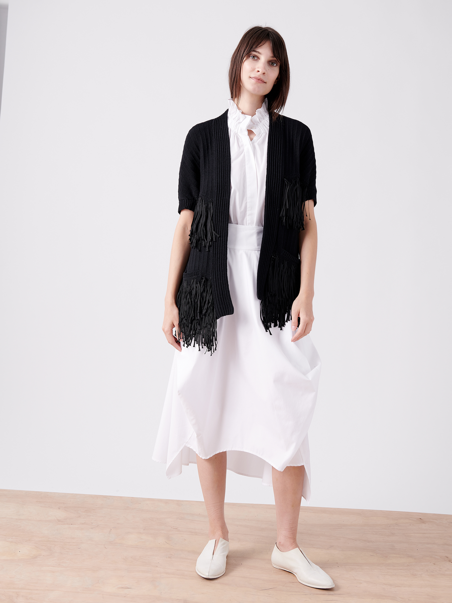 Dressed in a Fringe Hex Shrug from Zero + Maria Cornejo, crafted from organic cotton, a person with short dark hair stands on a wooden floor against a white backdrop. They wear a white dress and slip-on shoes, capturing the spirit of sustainable fashion.