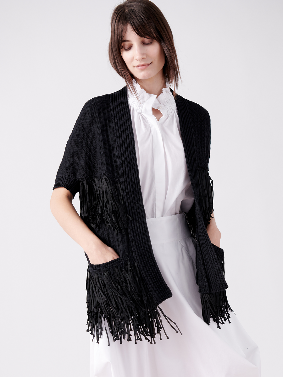 Donning the Fringe Hex Shrug by Zero + Maria Cornejo, a person wears a chic white dress and black fringe cardigan of organic cotton. Standing against a plain backdrop with short hair and hands in pockets, they embody a modern, sustainable style.