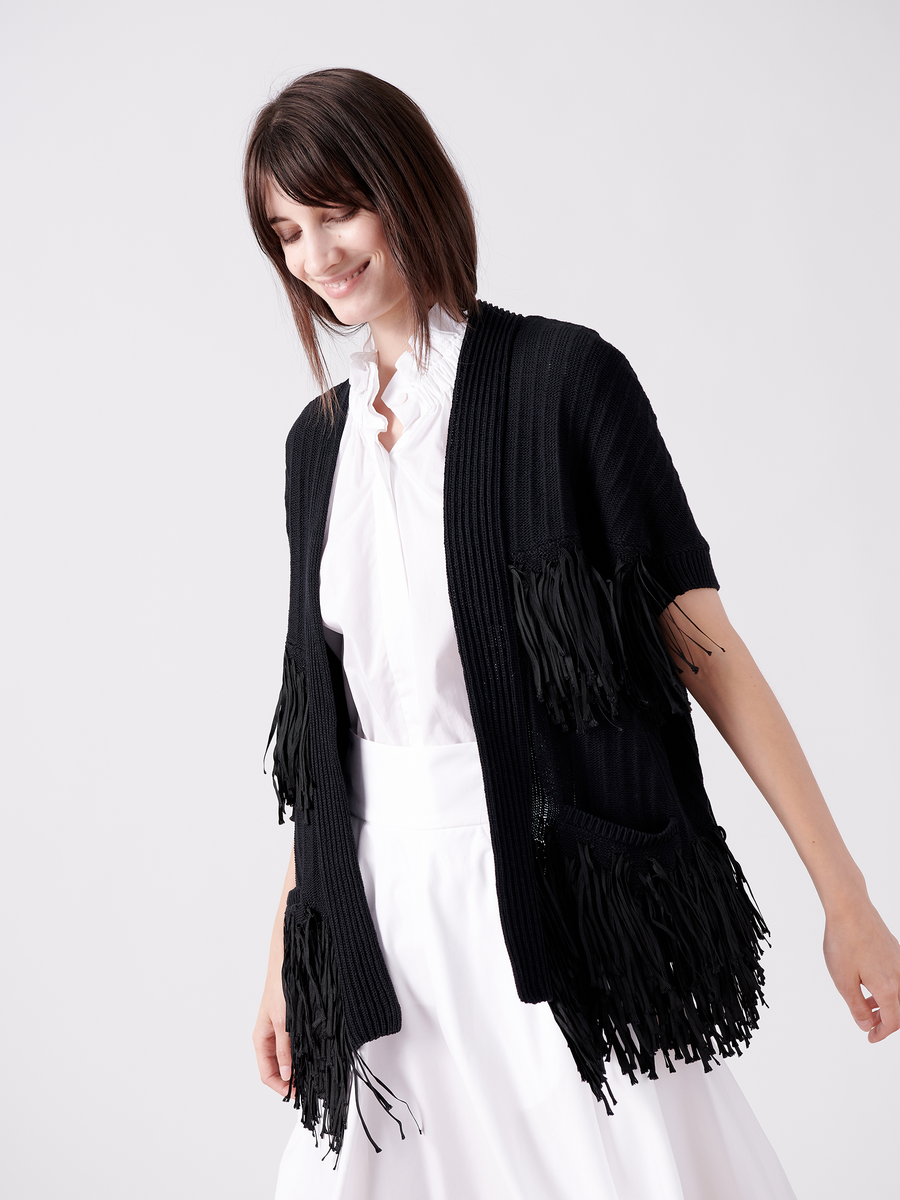 A woman with shoulder-length brown hair smiles, wearing Zero + Maria Cornejo's Fringe Hex Shrug in black over a white blouse and skirt. This ensemble, made from organic cotton, exemplifies sustainable fashion as she stands relaxed against a light gray backdrop.