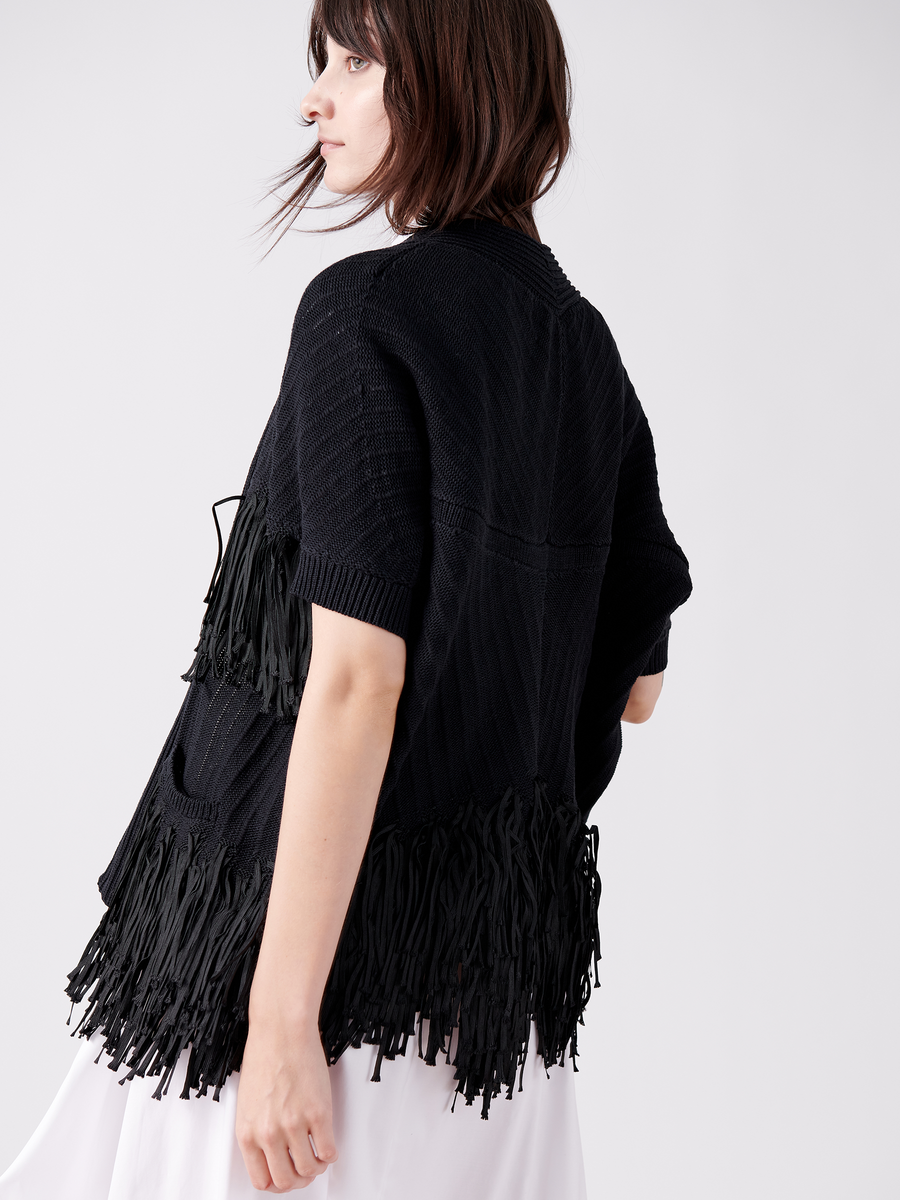 A person with shoulder-length brown hair wears the Fringe Hex Shrug by Zero + Maria Cornejo, a black, fringed shawl-style sweater over an organic cotton white outfit. Captured from the side against a plain white background, it's an elegant nod to sustainable fashion.