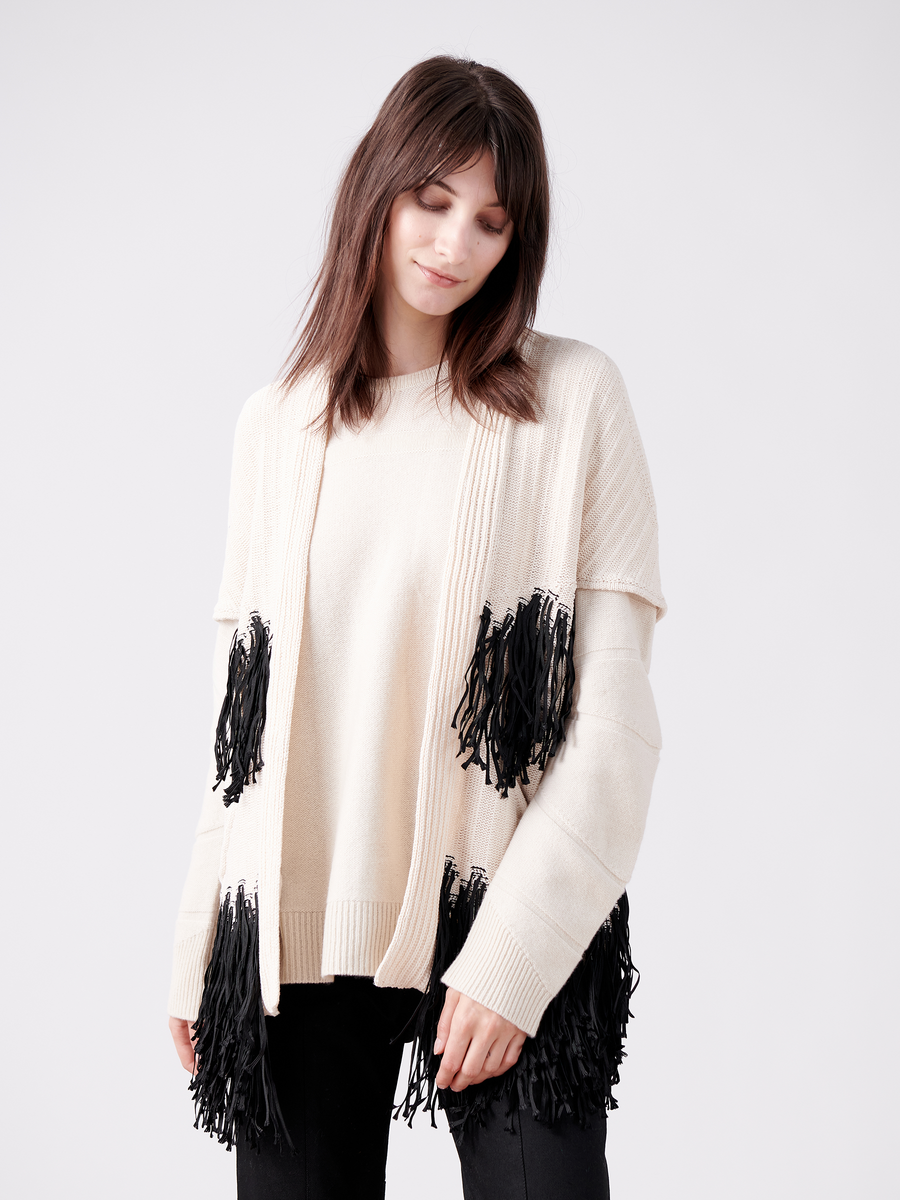 Against a plain white background, a woman embodies sustainable fashion in the Fringe Hex Shrug by Zero + Maria Cornejo. The cream sweater features black fringe details and is made from organic cotton. Her eyes are closed as her dark hair falls naturally around her shoulders.