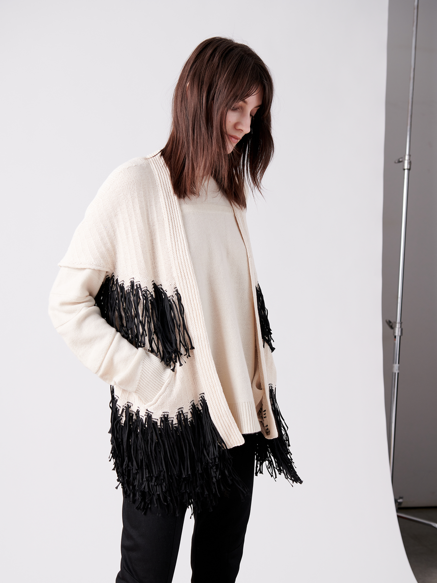 The woman wears the Fringe Hex Shrug by Zero + Maria Cornejo—a cream cardigan with black fringe, crafted from sustainable fashion materials. In a studio setting, her brown hair cascades gently as she looks down, elegantly paired with a neutral top and black pants.