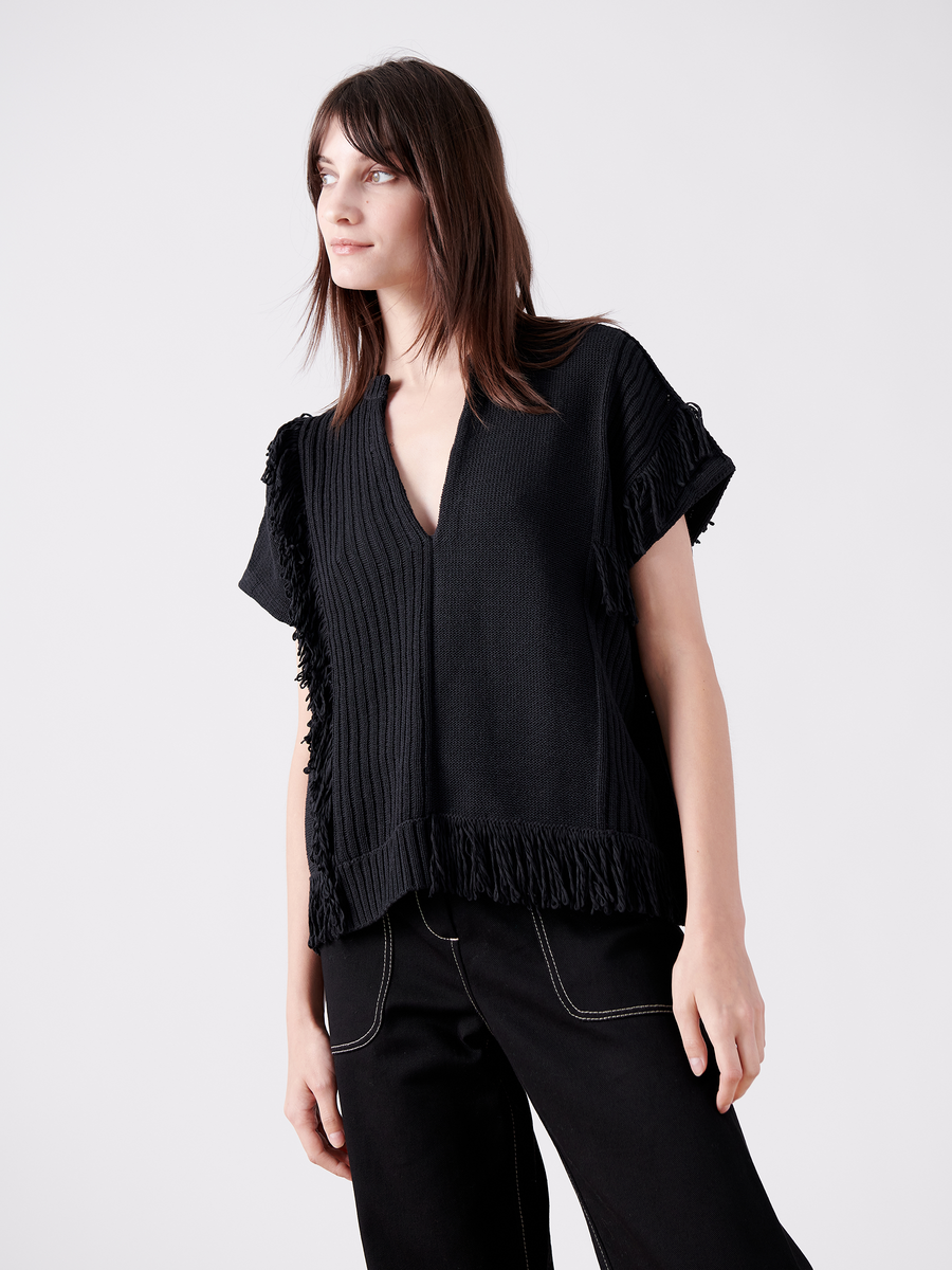 A person with shoulder-length brown hair wears the Patchwork Fringe Poncho by Zero + Maria Cornejo, styled in a loose black V-neck with fringe. Paired with black pants, they look slightly to the side against a plain white background.