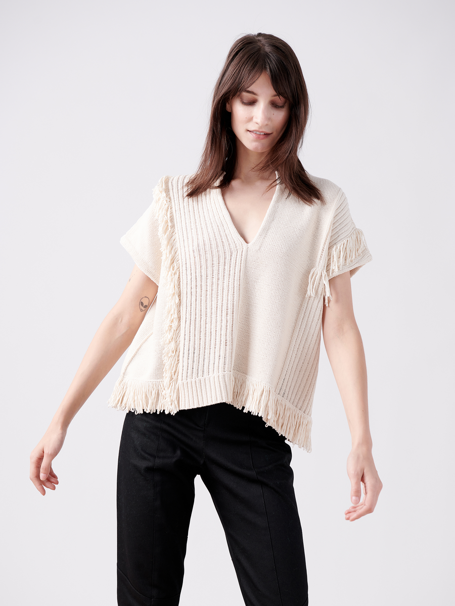 A person with long hair models the Patchwork Fringe Poncho by Zero + Maria Cornejo, featuring a cream hue and V-neck design. They pair it with black pants against a simple white backdrop, highlighting their dedication to sustainable fashion.