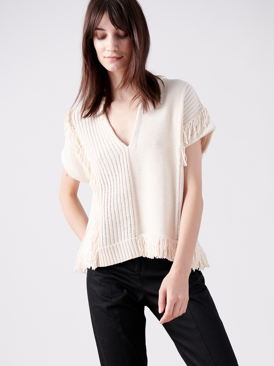 A person wears the Patchwork Fringe Poncho by Zero + Maria Cornejo, a light beige sustainable fashion top in cotton knit with V-neck and fringe-detailed sleeves and hem. Long brown hair falls as they stand against a plain light background, paired with black pants.