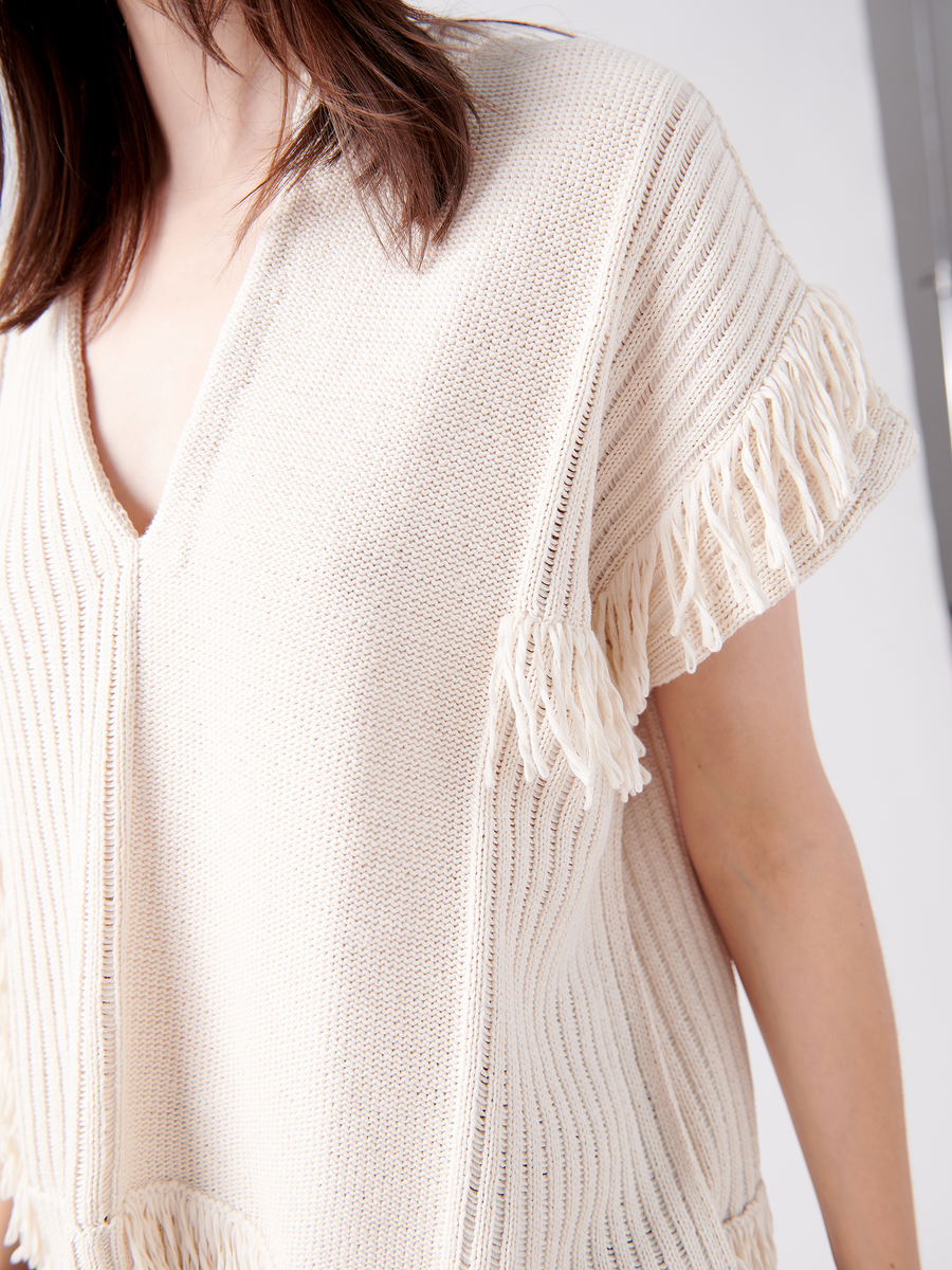 Against a neutral background, an unseen individual wears Zero + Maria Cornejo's Patchwork Fringe Poncho—crafted from cream-colored organic cotton with short sleeves and shoulder fringe—capturing the spirit of sustainable fashion.