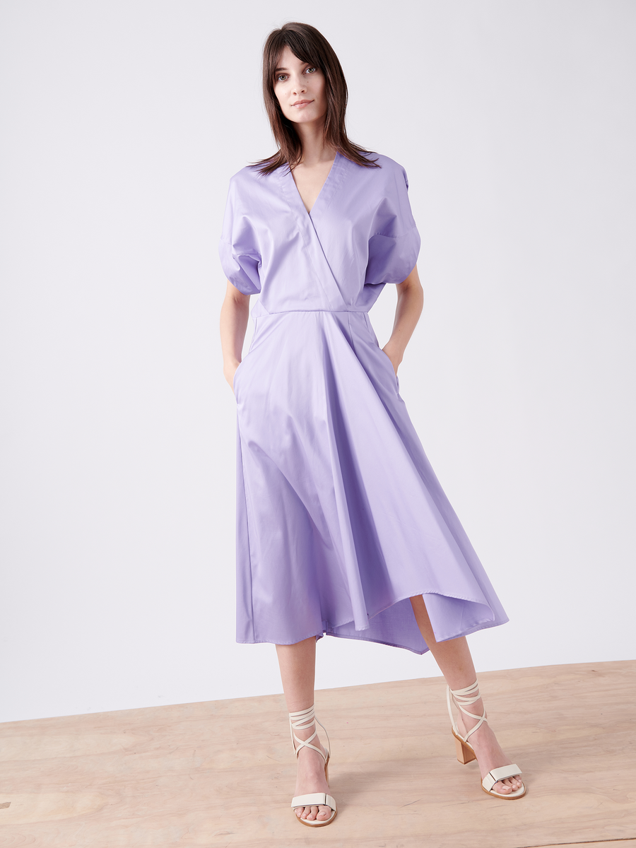 A model dons the Aki Wave Dress from Zero + Maria Cornejo, showcasing its wrap-effect design in lavender with a deep V-neck, puffed sleeves, and an asymmetrical skirt. Posing with hands in pockets on wooden flooring against a white backdrop, they're paired with strappy beige heels.