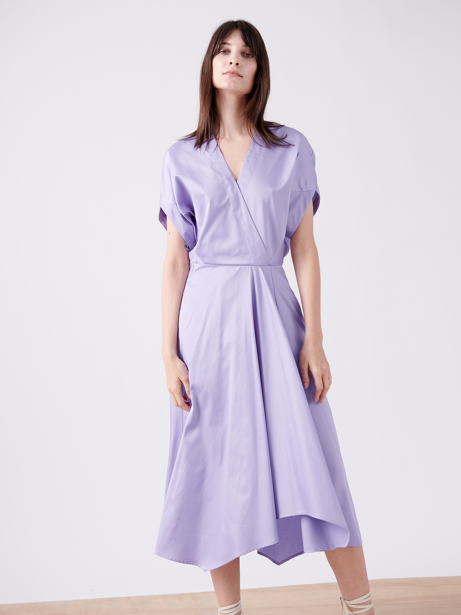 A person confidently wears the Aki Wave Dress by Zero + Maria Cornejo in light purple cotton broadcloth, featuring a wrap-effect V-neck, short sleeves, and a flowing asymmetrical hem. They pose indoors against a plain white background with long, dark hair.