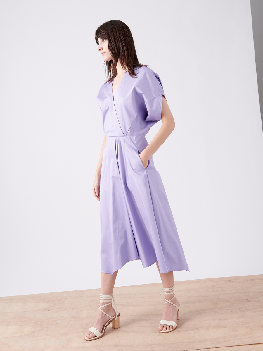 A woman in a Zero + Maria Cornejo Aki Wave Dress gazes to the side. This lavender, wrap-effect, cotton broadcloth dress features a loose fit, short sleeves, and pockets. Her straight hair and strappy white high-heeled sandals complete the look against a plain background.