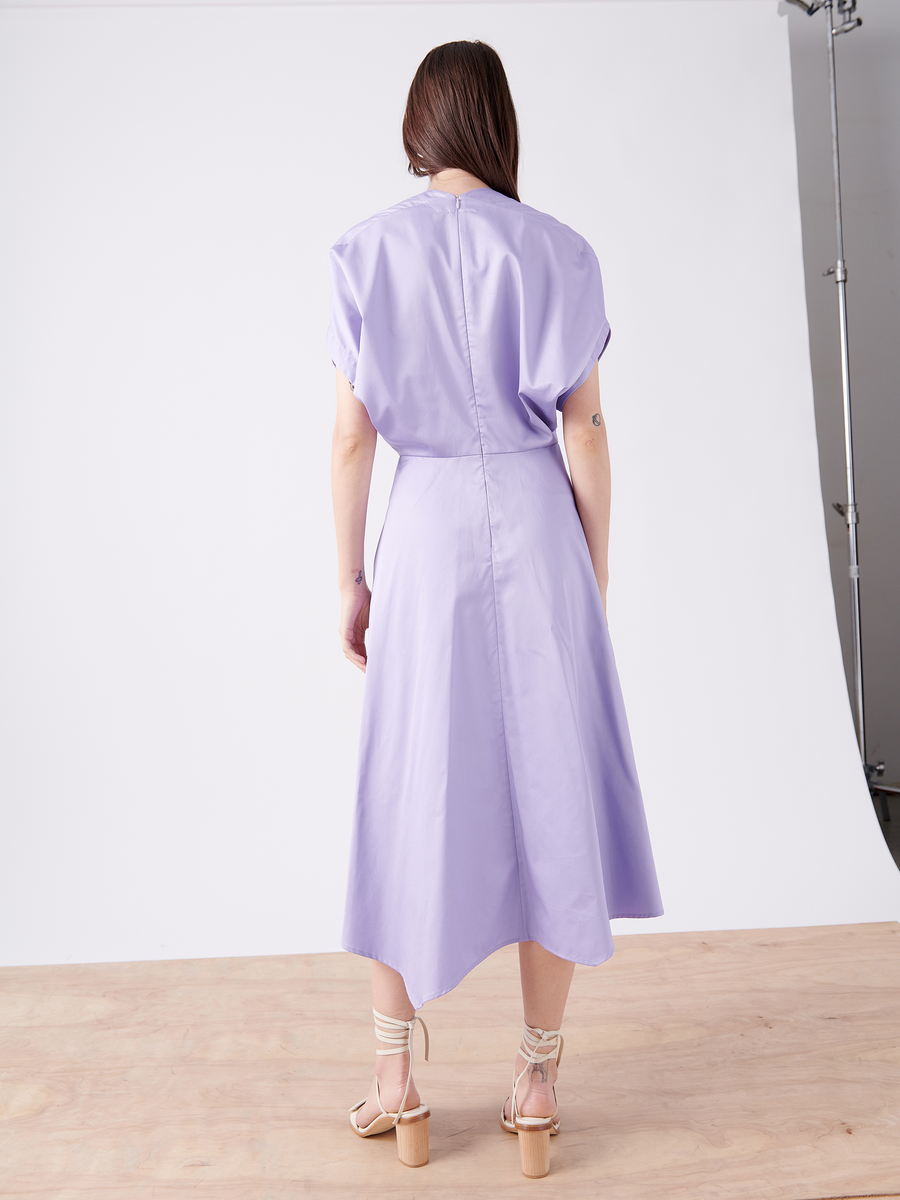 A person with long hair stands back to the camera in a lavender Aki Wave Dress by Zero + Maria Cornejo, featuring short sleeves and a mid-calf hem. They elegantly pair it with beige heeled sandals on a wooden floor, set against a pristine white background.