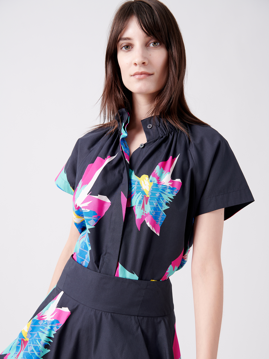 A person with long brown hair wears the Zero + Maria Cornejo Elastic Gaban Shirt, boasting vivid abstract floral patterns in pink, blue, and teal on a dark, short-sleeved dress. It features an elastic stand collar and fitted waist against a plain light background.