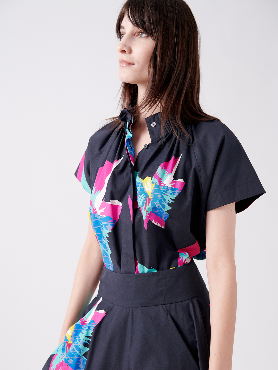 A woman stands gracefully, wearing a Zero + Maria Cornejo Elastic Gaban Shirt with vibrant blue, pink, and white floral prints on cotton. Her long dark hair flows as she gazes to the side calmly. The shirt features a short-sleeve design and elegant elastic stand collar on a light-colored background.