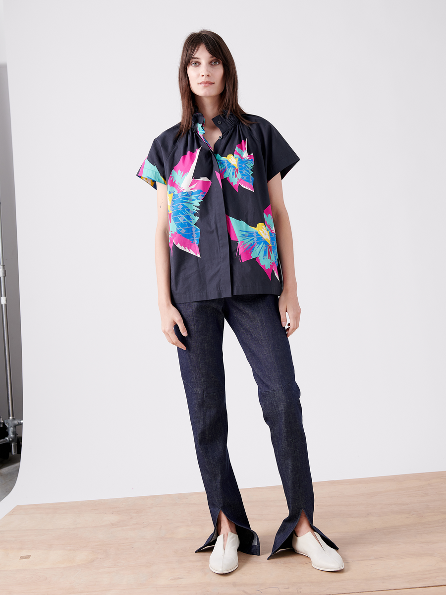 A woman stands on a wooden platform against a plain backdrop, wearing Zero + Maria Cornejo's Elastic Gaban Shirt with colorful floral patterns and short sleeves, paired with dark split-hem pants. Her white shoes contrast with the outfit. She has a neutral expression and wears her hair down.