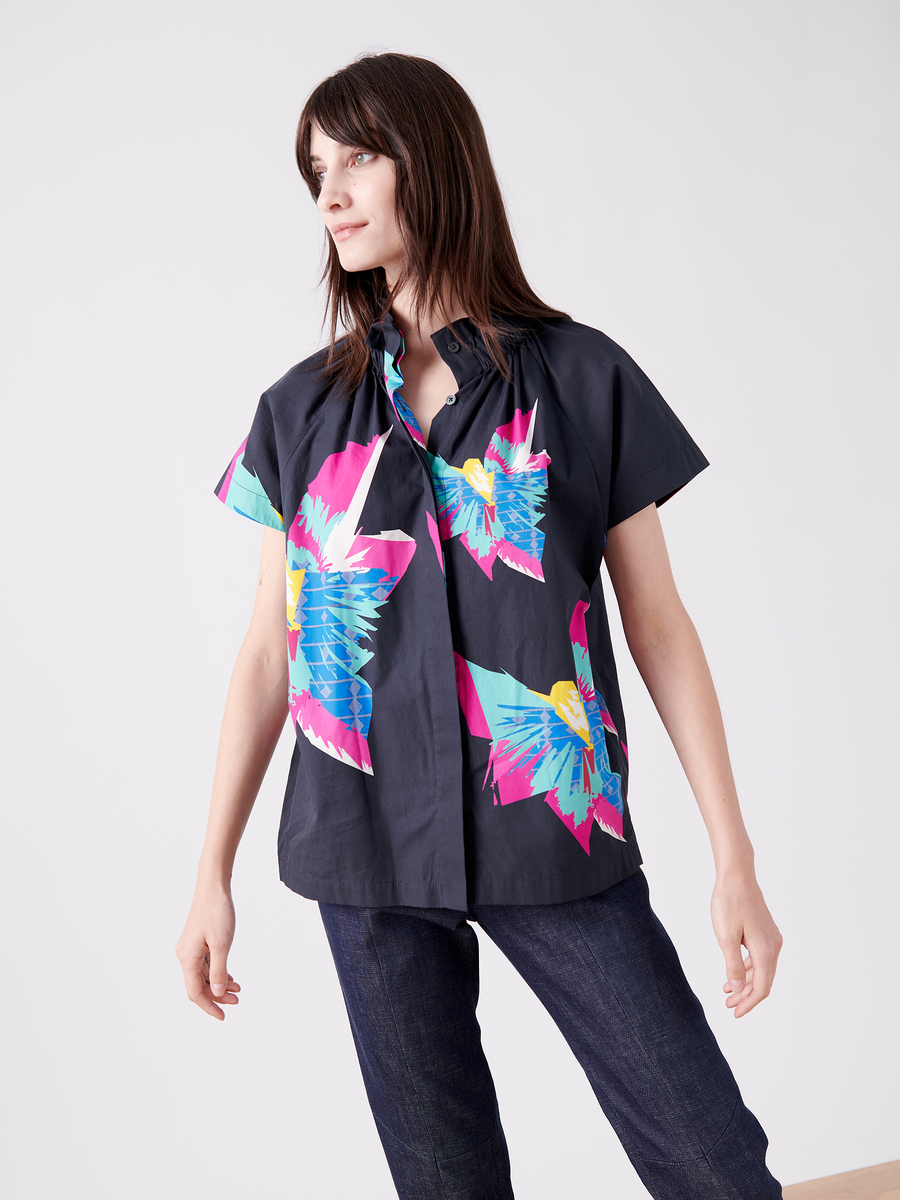 A person with long brown hair wears the Zero + Maria Cornejo Elastic Gaban Shirt with bold pink and blue floral designs on a black base, paired with dark jeans. The shirt features an elastic stand collar for style and comfort against a plain light gray background.