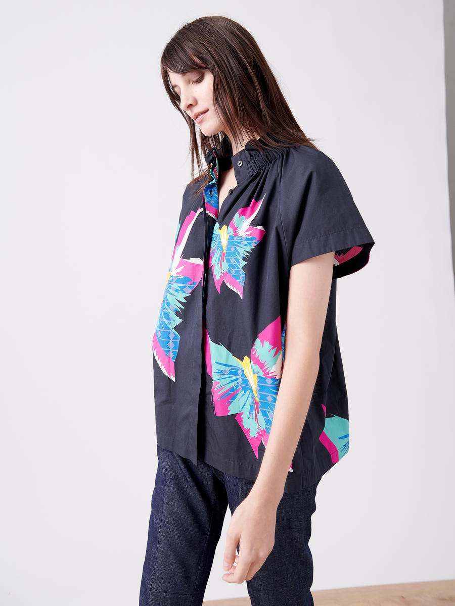 A person with long hair in the Elastic Gaban Shirt by Zero + Maria Cornejo stands against a plain backdrop. The dark, colorful butterfly-patterned shirt boasts an elastic stand collar for elegance, paired with a loose fit and short sleeves. They gaze slightly downward.