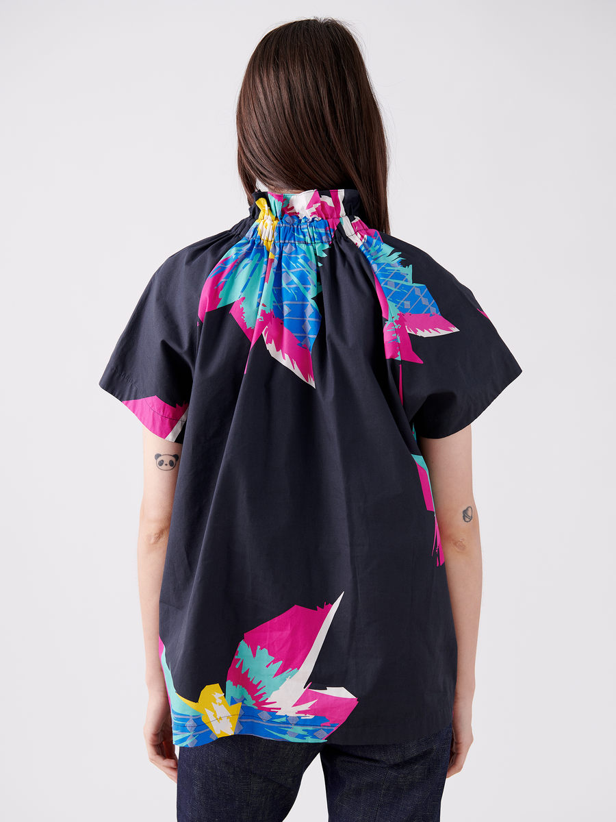 Seen from behind, a person with long dark hair wears the short-sleeve Elastic Gaban Shirt by Zero + Maria Cornejo, featuring abstract floral patterns in pink, blue, and yellow. The cotton shirt complements their small tattoos and pairs effortlessly with dark jeans. Final Sale.