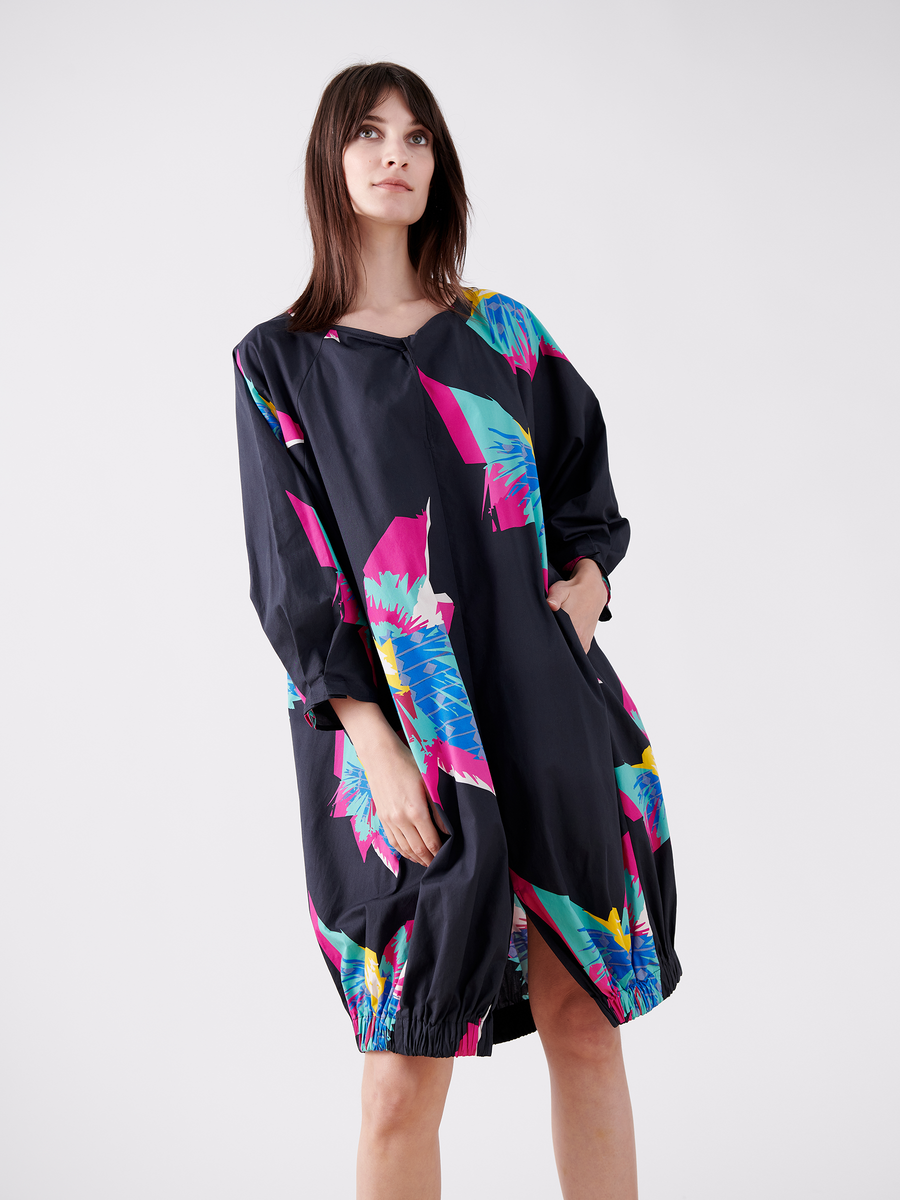 A woman with long brown hair stands against a plain background, hands in the pockets of her colorful Long-Sleeved Luca Dress by Zero + Maria Cornejo, featuring abstract pink, blue, and green patterns.