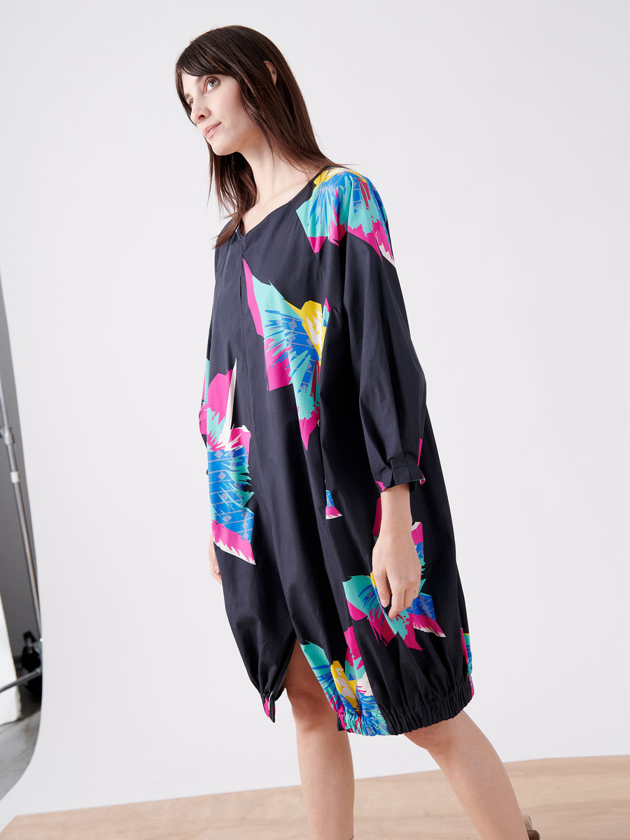 A person in a knee-length, printed Long-Sleeved Luca Dress by Zero + Maria Cornejo, boasting vibrant abstract patterns of blue, pink, and yellow on dark cotton, stands against a plain white background. The tropical print harmonizes with their long dark hair as they gaze off to the side.