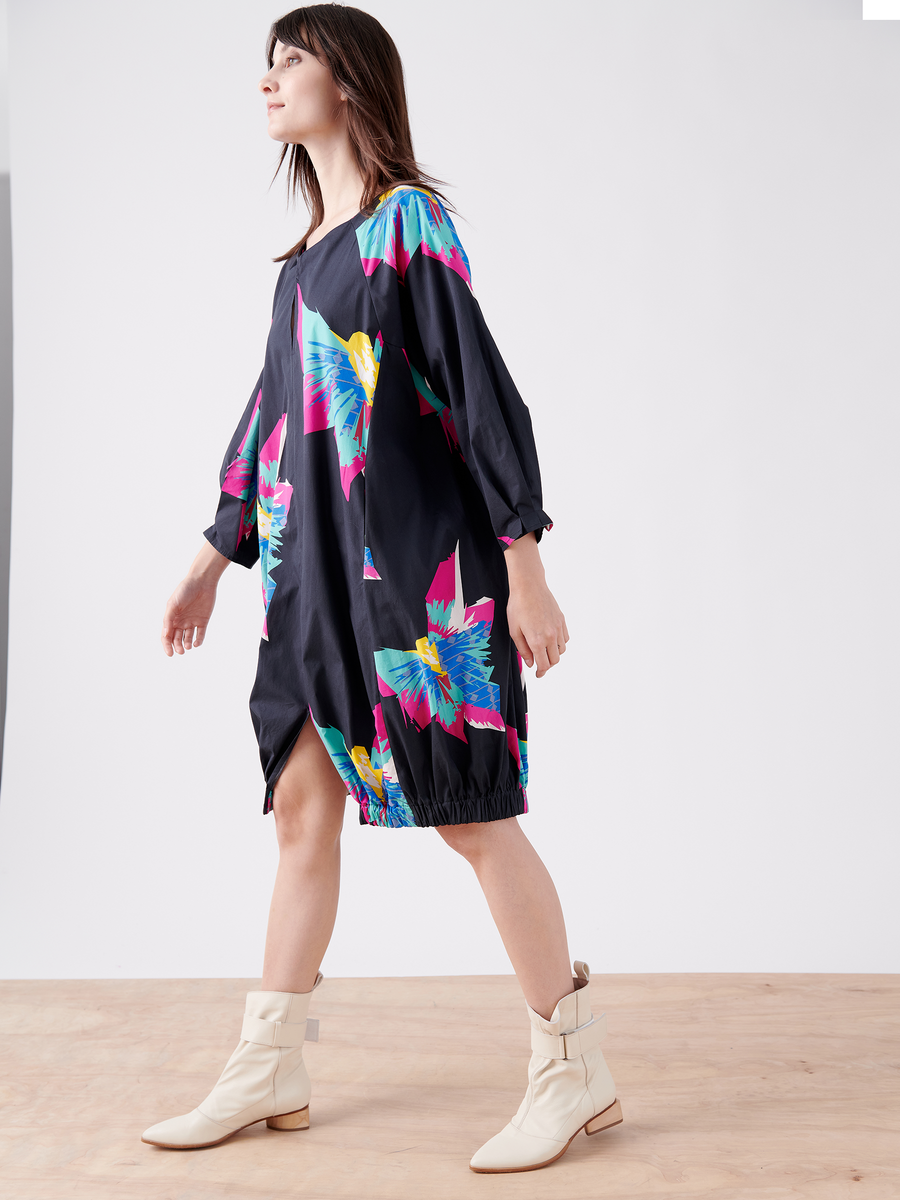 Someone in a Long-Sleeved Luca Dress by Zero + Maria Cornejo with a vibrant tropical print walks on a wooden floor against a plain white background. They wear light-colored ankle boots and have long dark hair.