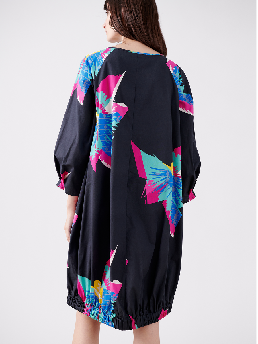 A person models the Long-Sleeved Luca Dress by Zero + Maria Cornejo, showcasing its loose fit and abstract butterfly pattern in blue, pink, and white on a dark backdrop. The elastic hem and draped silhouette stand out against a plain white background.