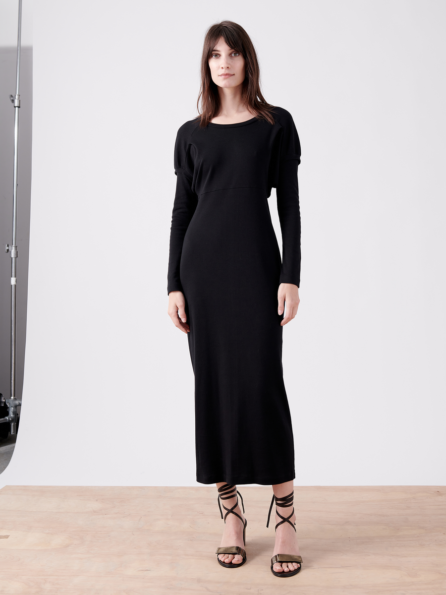 A woman, with long dark hair, stands on a wooden floor and plain white background, wearing the Zero + Maria Cornejo Rib Long-Sleeved Ama Dress—Final Sale and strappy high-heeled sandals. She gazes directly at the camera.