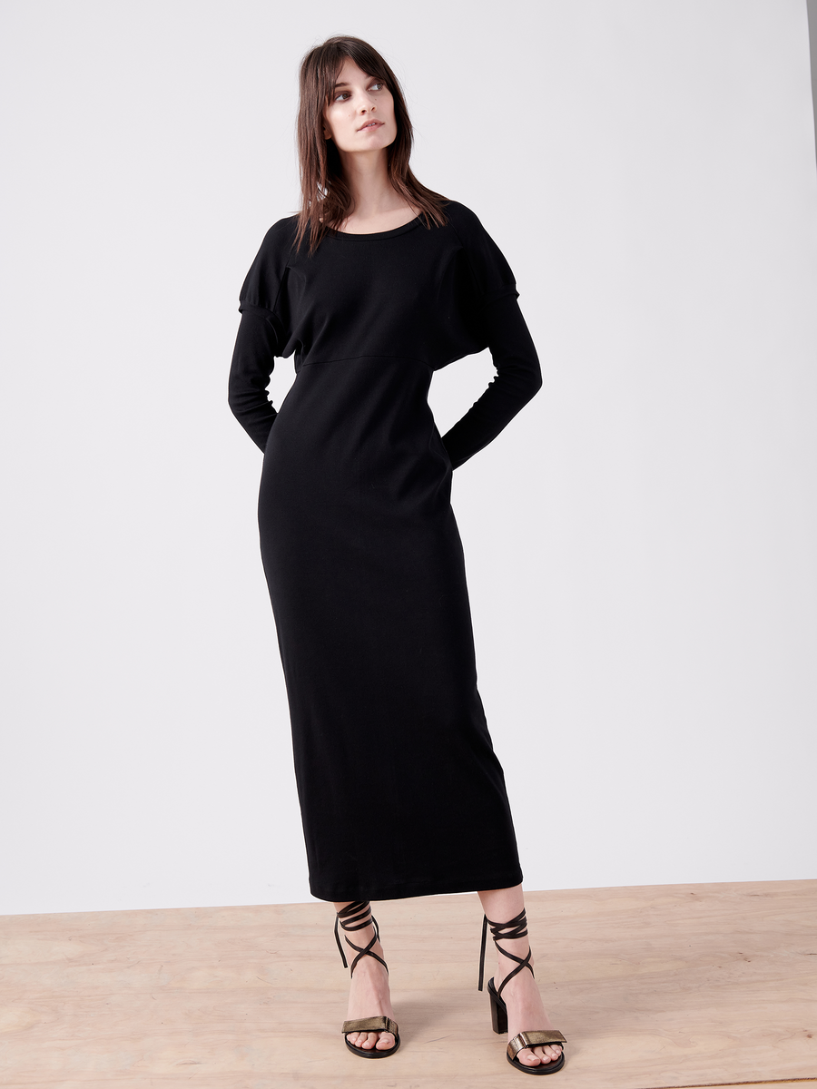 A woman stands on a wooden surface against a plain background wearing the Rib Long-Sleeved Ama Dress by Zero + Maria Cornejo, featuring ruffle sleeve details and an oversized silhouette. Her arms are behind her back, and she complements the look with strappy black high-heeled sandals.