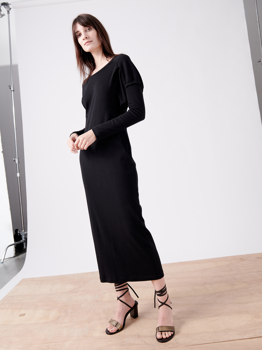 A woman confidently poses on a wooden platform in the Rib Long-Sleeved Ama Dress by Zero + Maria Cornejo and strappy high-heeled sandals, against a plain white backdrop with studio lights and equipment partially visible.