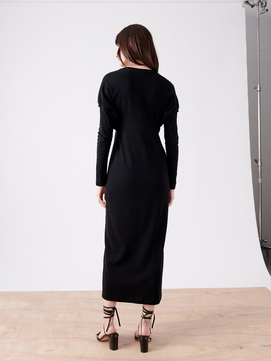 A woman with long brown hair stands on a wooden floor, wearing Zero + Maria Cornejo's Rib Long-Sleeved Ama Dress - Final Sale. She faces away from the camera in strappy high heels, with the oversized silhouette enhancing the plain white studio setup.