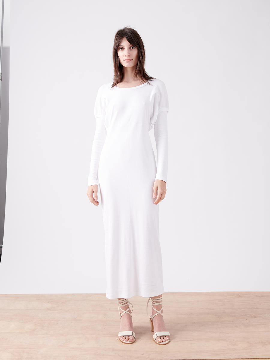 A woman stands on a wooden floor against a plain white background, wearing the oversized Rib Long-Sleeved Ama Dress by Zero + Maria Cornejo, crafted from Japanese cotton, matched with strappy high-heeled sandals. Her shoulder-length hair frames her face as she gazes forward neutrally.