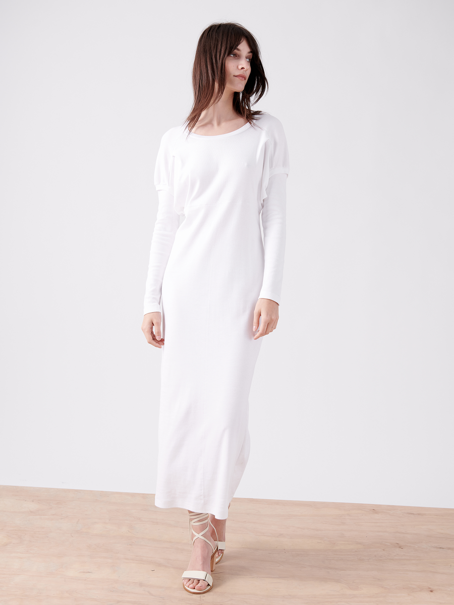 On a wooden floor, a woman with long dark hair looks to the side in an oversized silhouette. She wears the Rib Long-Sleeved Ama Dress by Zero + Maria Cornejo, paired with strappy high-heeled sandals against a plain white background.