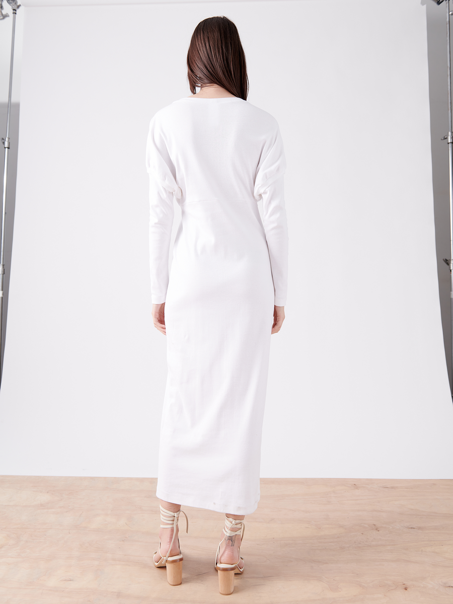 A woman with long brown hair faces away, wearing a Rib Long-Sleeved Ama Dress by Zero + Maria Cornejo and beige heels. The oversized silhouette complements the studio's plain white backdrop and wooden floor.