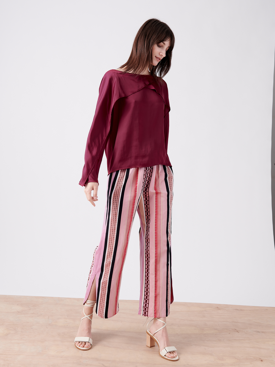 A woman stands on a wooden floor, wearing the Zero + Maria Cornejo Ido Shirt, a fluttery long-sleeved burgundy blouse, and pink pants with vertical black and red stripes. Her brown hair cascades downward as she looks down, anchored by beige heeled sandals.