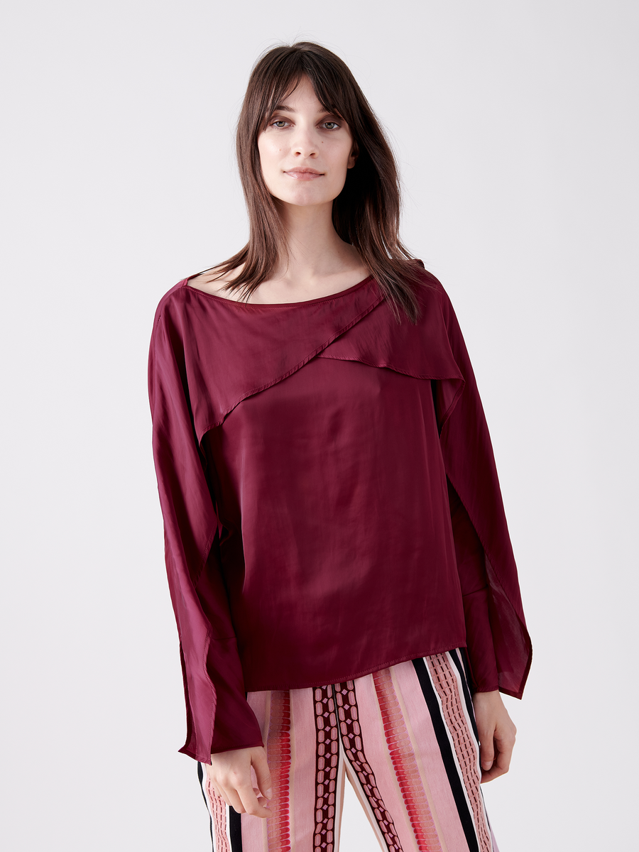 A woman with long dark hair wears the Ido Shirt by Zero + Maria Cornejo. The loose, burgundy satin blouse features fluttery long sleeves, adding elegance as she stands in colorful striped pants against a plain light gray background with a relaxed expression.