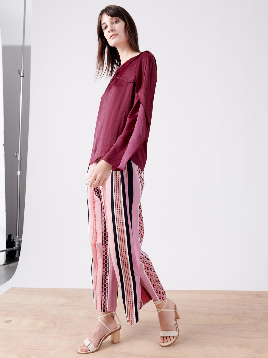 A woman poses on a wooden platform in an Ido Shirt by Zero + Maria Cornejo, styled with fluttery long sleeves. She's wearing patterned pink pants with vertical stripes, beige high-heeled sandals, and has long brown hair against a white studio backdrop.