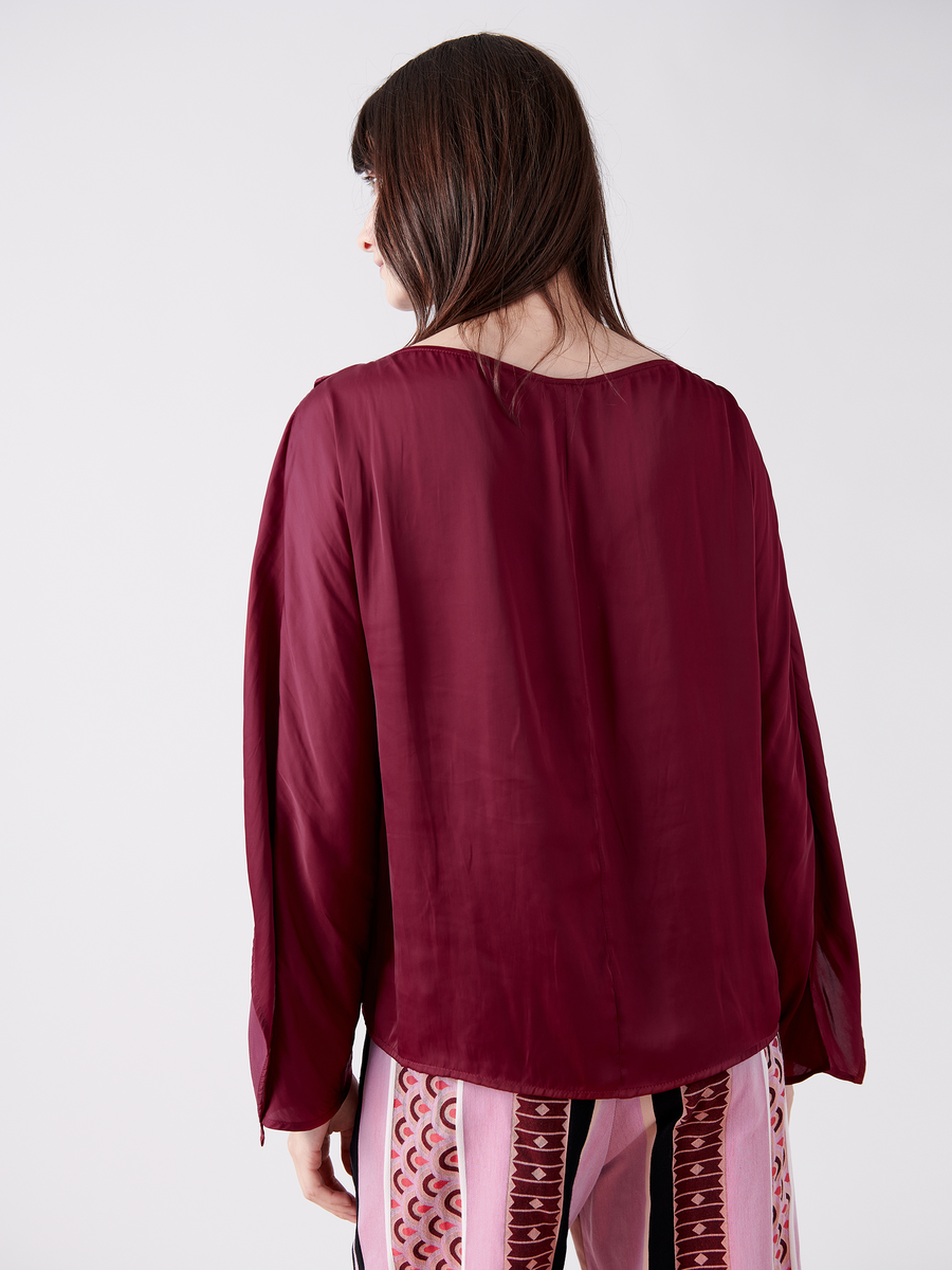 A person with long dark hair, facing away, wears the Ido Shirt from Zero + Maria Cornejo. This washed georgette shirt has fluttery long sleeves in maroon paired with beige pants featuring a red geometric pattern, set against a plain white background. Made in New York.