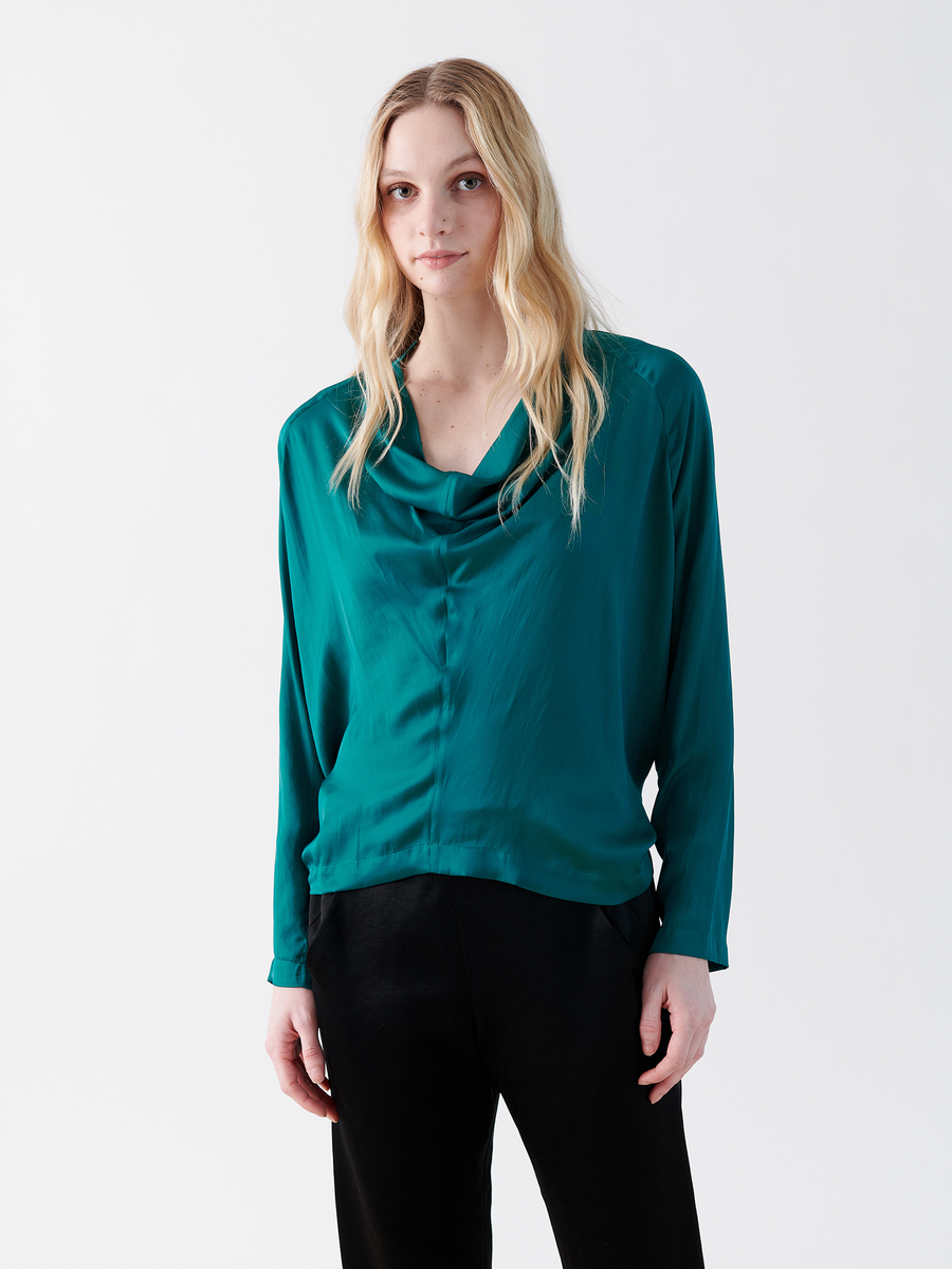 A person with long, wavy blonde hair stands against a white background, wearing the branded Long-Sleeved Evi Top by Zero + Maria Cornejo, featuring a teal washed georgette and a draped open neckline, paired with black pants. Their expression is neutral, with hands casually in pockets.