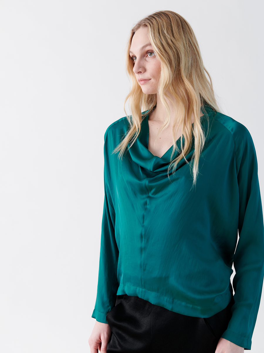 A person with long blond hair stands against a plain white background, wearing the Long-Sleeved Evi Top by Zero + Maria Cornejo, featuring a draped open neckline in washable teal polyester as they gaze to the side with a neutral expression.