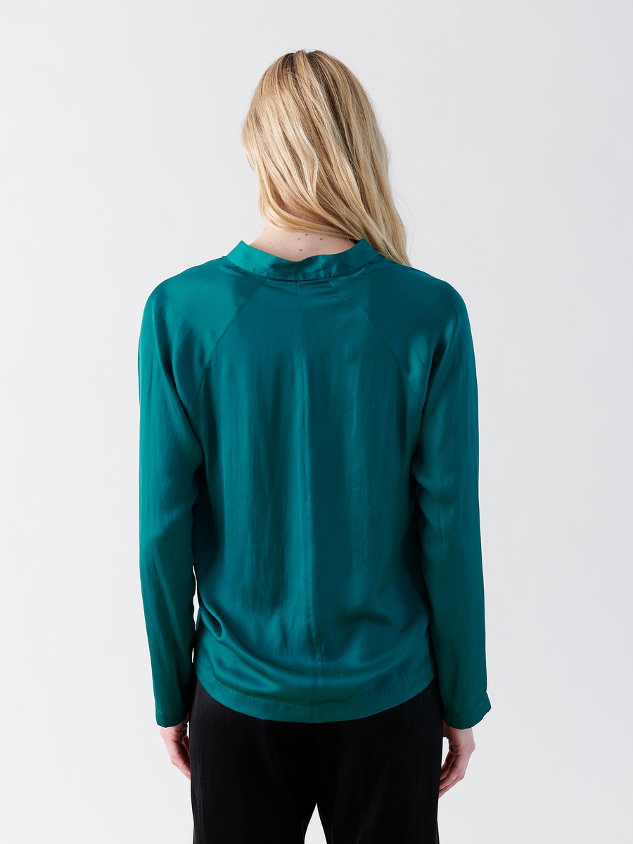 A person with long blonde hair faces away from the camera, wearing a teal washed Long-Sleeved Evi Top by Zero + Maria Cornejo with a draped open neckline and black pants against a plain white background.