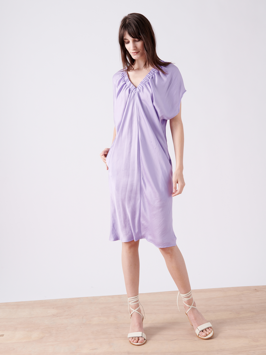 A person with long brown hair wears the Ruched Tilly Dress by Zero + Maria Cornejo, a light purple knee-length dress with a gathered neckline made of washable fabric. They stand on a wooden floor against a plain white background, paired with strappy white sandals.