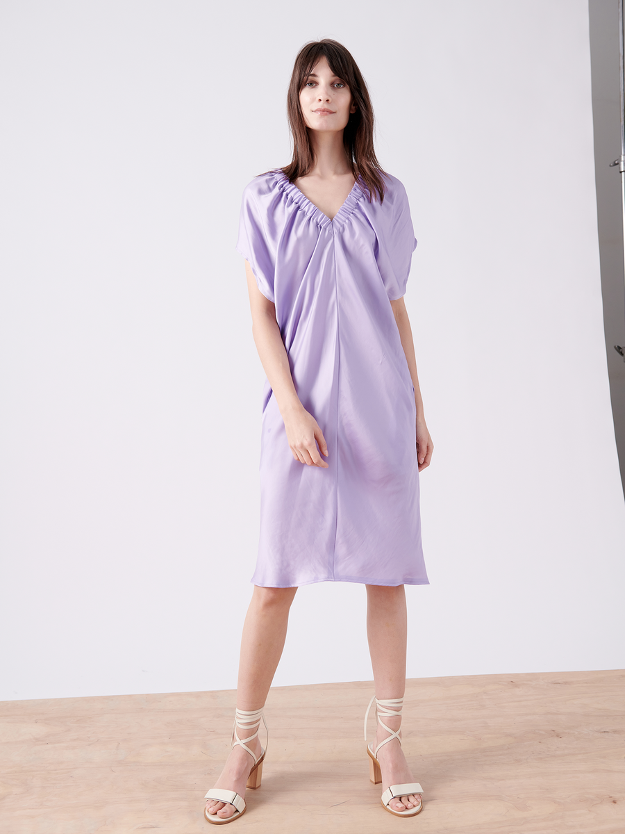 A woman poses elegantly in the Ruched Tilly Dress by Zero + Maria Cornejo, a knee-length lavender piece with a gathered neckline. With medium-length dark hair and white strappy heels, she stands against a plain white studio backdrop.