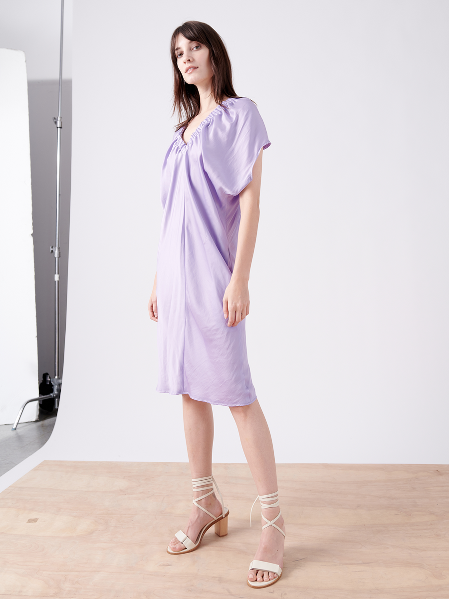 A woman in a light purple Ruched Tilly Dress by Zero + Maria Cornejo stands on a wooden floor against a plain white background. She wears strappy beige sandals and has straight, shoulder-length hair. The knee-length dress is washable with short sleeves and a gathered neckline.