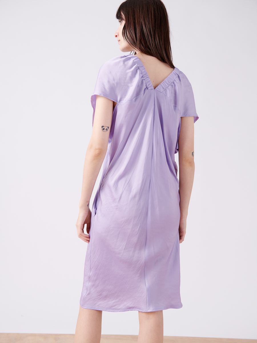 A person with long dark hair stands facing away, wearing the silky Ruched Tilly Dress - Final Sale by Zero + Maria Cornejo. This knee-length lavender dress has short sleeves and a gathered V-shaped back, crafted from washable fabric. Small tattoos adorn their upper arms against a softly lit plain background.