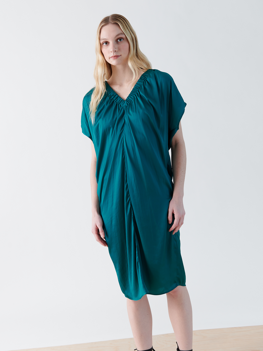 A person with long blonde hair models the Ruched Tilly Dress by Zero + Maria Cornejo, a teal, knee-length dress with short sleeves and a V-neck. The dress has a gathered design and is made from washable fabric, set against a plain white background.