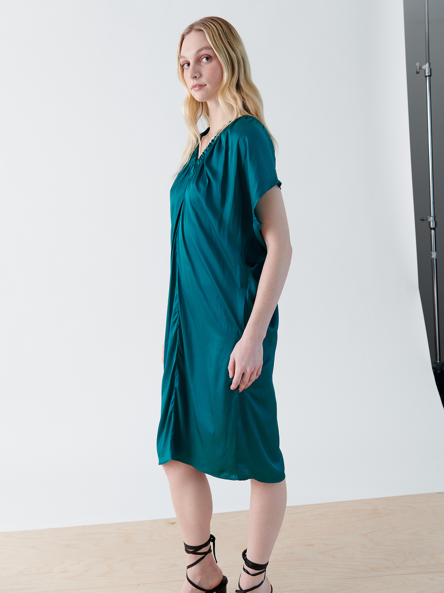 A woman with long blonde hair poses in a studio wearing the Ruched Tilly Dress from Zero + Maria Cornejo, a teal knee-length washable dress with a v-neck. The elegant ensemble pairs perfectly with her black strappy heels against the white background and light wooden floor.