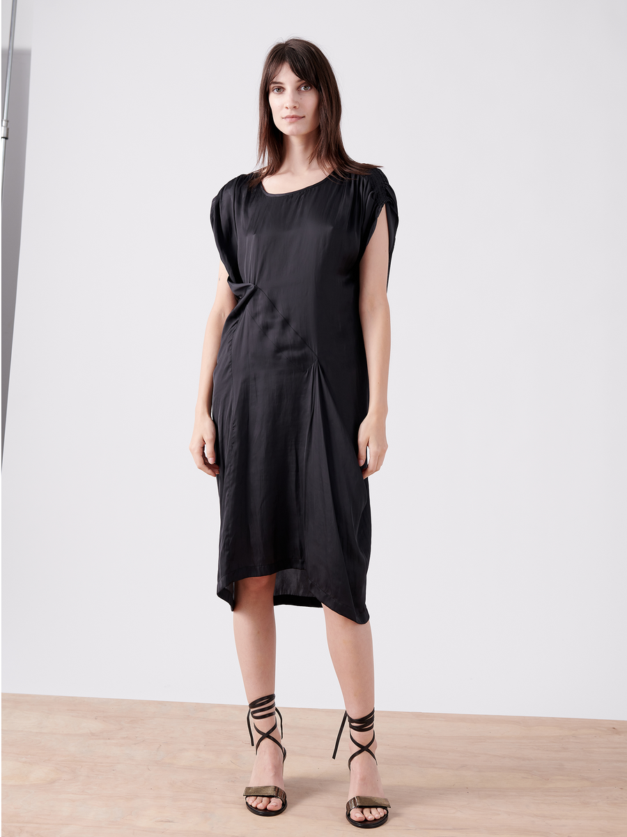 A woman stands on a wooden floor wearing the Ruched Fold Dress - Final Sale by Zero + Maria Cornejo. This black, short-sleeve, knee-length washed georgette dress features an asymmetrical folded front. She has long dark hair and wears lace-up high heels against a light gray backdrop.