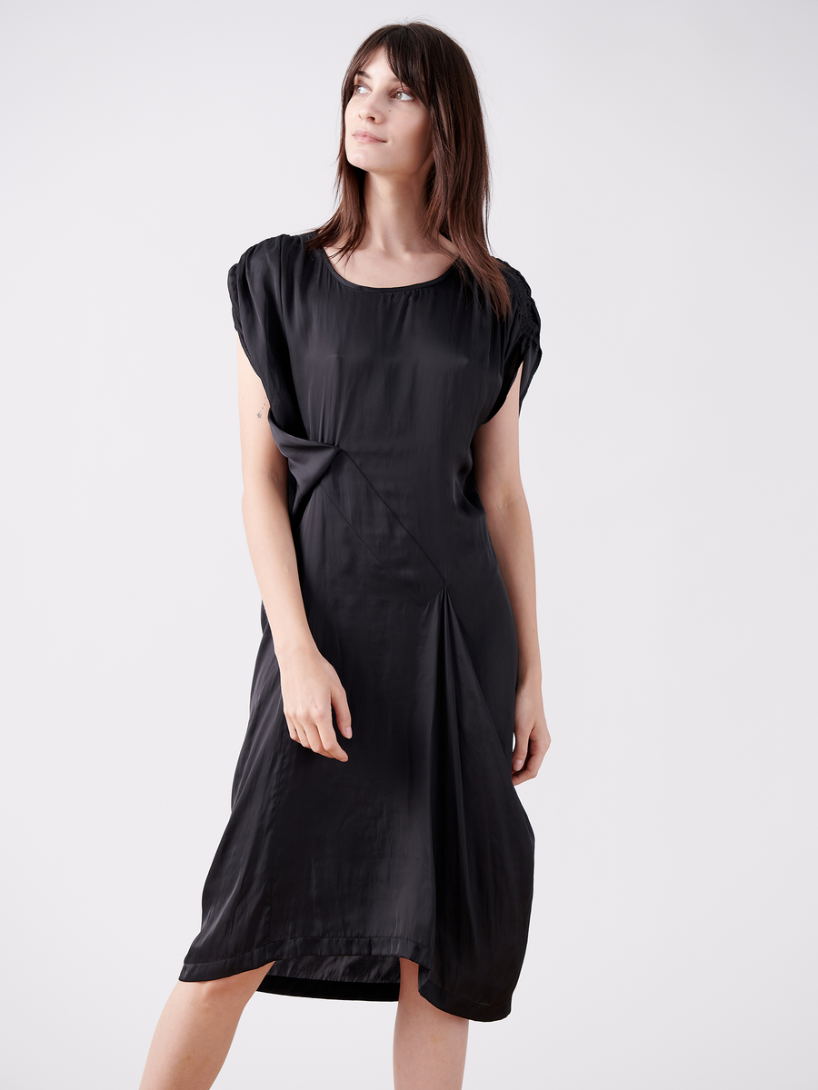 A woman in a Ruched Fold Dress from Zero + Maria Cornejo's final sale stands against a plain white background. The black washed georgette dress features an asymmetrical folded front with cap sleeves. She has straight brown hair and gazes slightly upward with a relaxed expression.