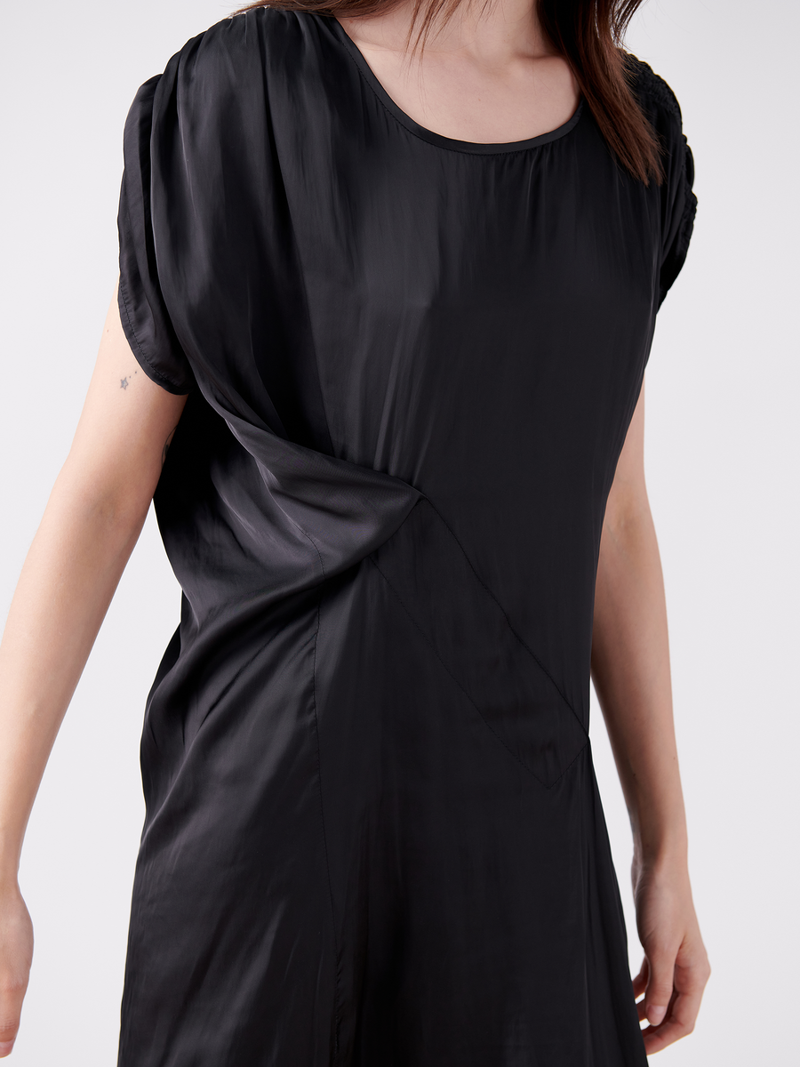 A person with long hair is wearing the Ruched Fold Dress - Final Sale by Zero + Maria Cornejo. This loose-fitting, short-sleeved black dress features a round neckline, washable fabric with subtle sheen, and gathered detail on one side against a plain, light-colored background.